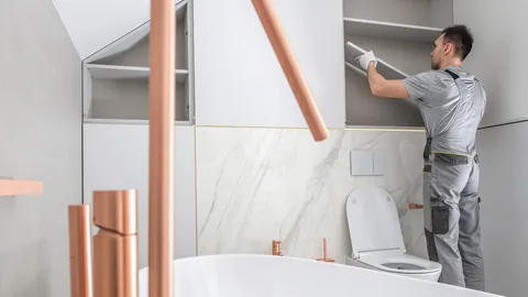 Professional carpenter installing sleek white custom bathroom storage cabinets above a luxury bathtub with copper fixtures.
