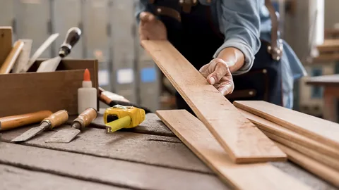 Close-up of a carpenter's hands selecting premium wood boards alongside a tape measure and chisels to craft custom bathroom furniture.